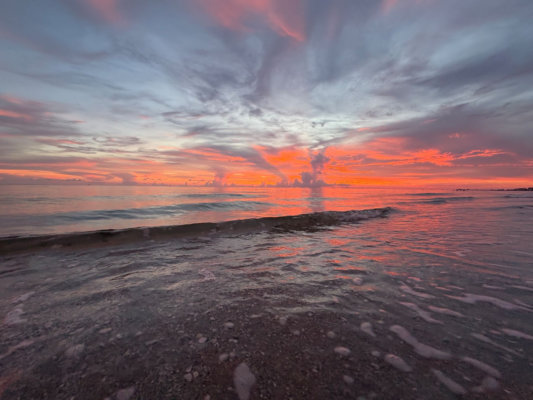 A vibrant sunset casts colorful reflections over gentle ocean waves approaching the shore.