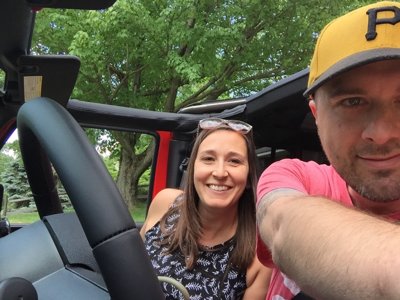 A smiling woman and a man wearing a yellow cap are sitting inside a car with the top down, surrounded by green trees.